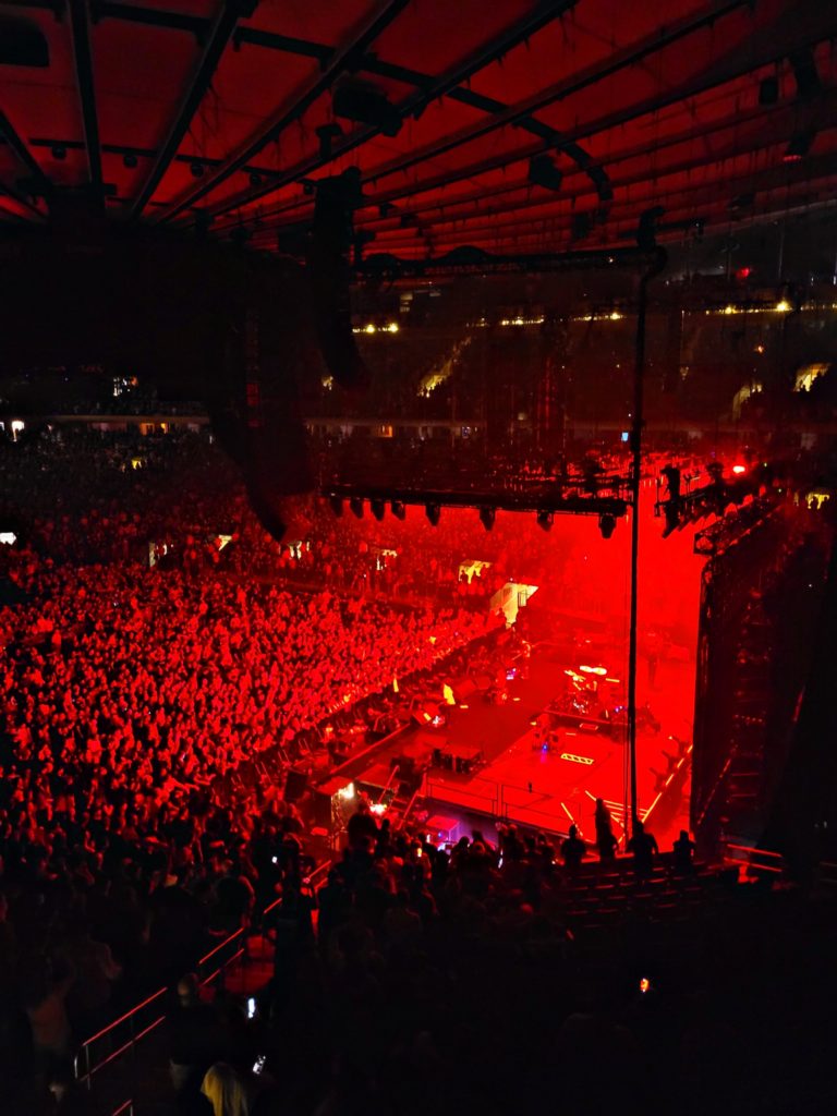 Red and black hues of a sold out concert at Madison Square Garden. The red is coming from stage lights above that cast a glow over the audience of hundreds of bodies.