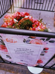 Inside a barricade is a bouquet of Pink, red, and yellow flowers. A flowery sign reads, "Join Us for a Candlelight Vigil Honoring Those Lost to Covid-19"