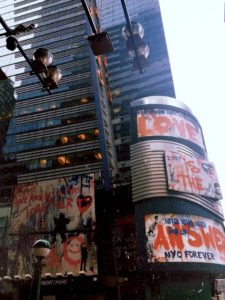 Two graffiti-like billboards in Times Square. Red letters read phrases, "Love is the Answer," "Nyc Forever," "We are all in this together," and "Never, Never, Never Give up!"