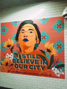 A subway ad with a woman is on the center with blue lipstick. Bold words underneath her read, "I still believe in our city."