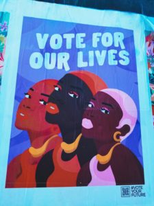 A poster with three Black woman looking in the same direction. Lettered in white is the text, "Vote for our lives."